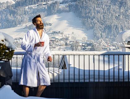 Hotelgast im Bademantel steht auf einem schneebedeckten Balkon in der Sonne 
