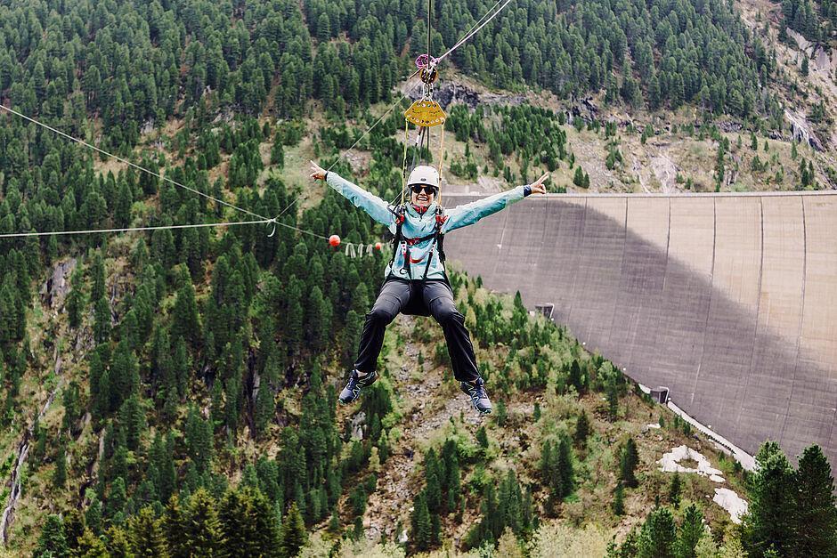 Eine Frau hängt an der Flying Fox Bahn Schlegeis 131 hoch über dem Zillertal und strahlt in die Kamera