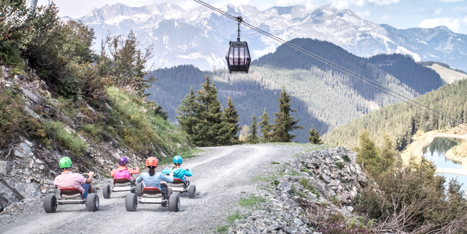 Eine Familie fährt vor der Bergkulisse und Gondel auf Carts den Berg hinunter
