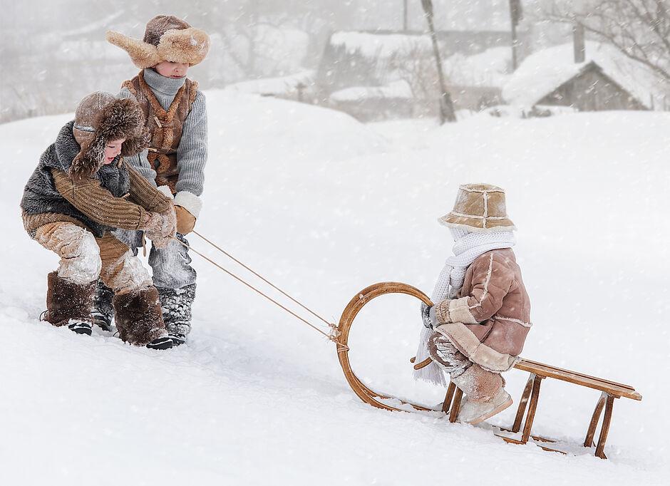 Zwei Kinder ziehen ein drittes Kind auf einem Rodel einen kleinen schneebedeckten Hang hinau