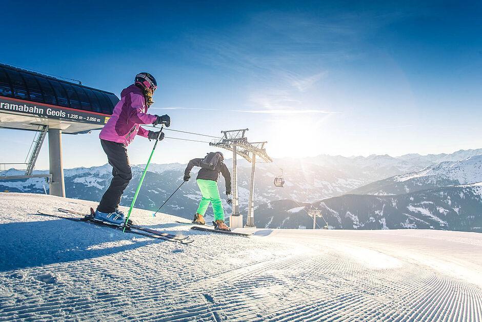 Im Winterurlaub in Tirol beginnen zwei Skifahrer an einer modernen Bergbahn ihre Abfahrt und schauen dabei in Richtung des grandiosen Alpenpanoramas