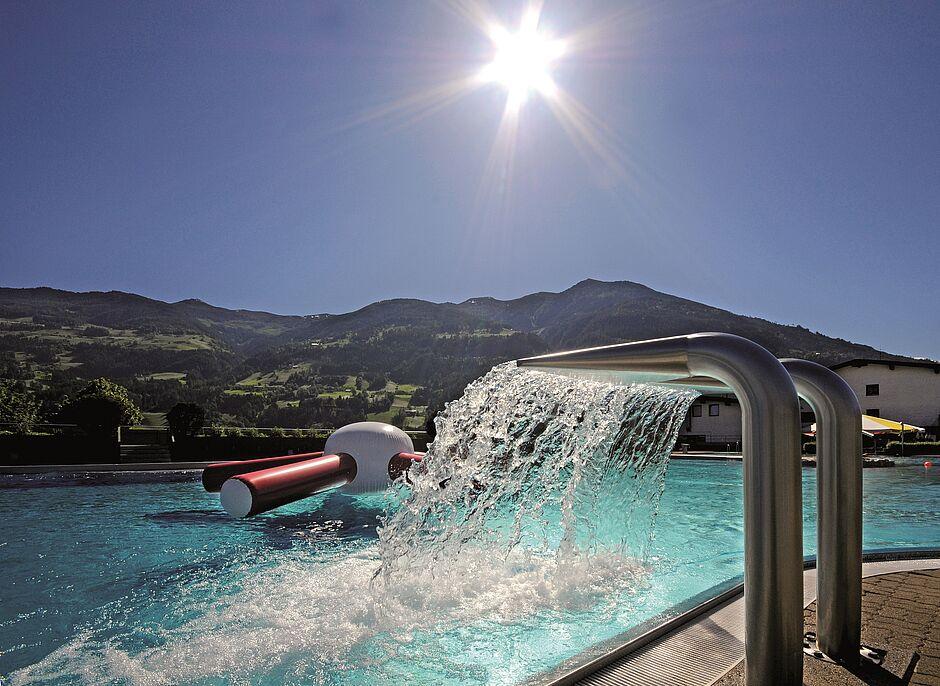 Die Sonne steht zur Sommerurlaubs-Saison in Tirol hoch am Himmel und scheint auf einen klaren Pool mit Bergpanorama
