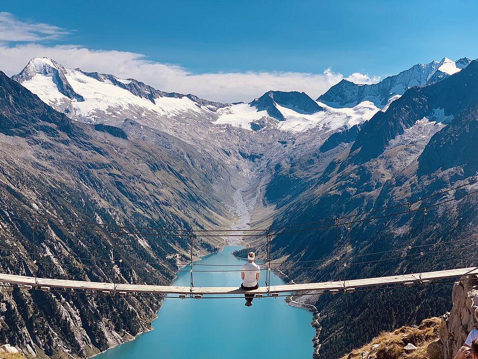 Mann sitzt auf einer hohen Hängebrücke über einem See in den Bergen 