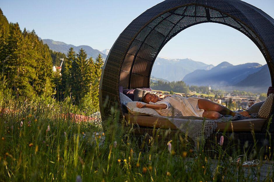 Eine Frau liegt in einem Rattanpavillion inmitten einer Wiese mit den Zillertaler Bergen im Hintergrund