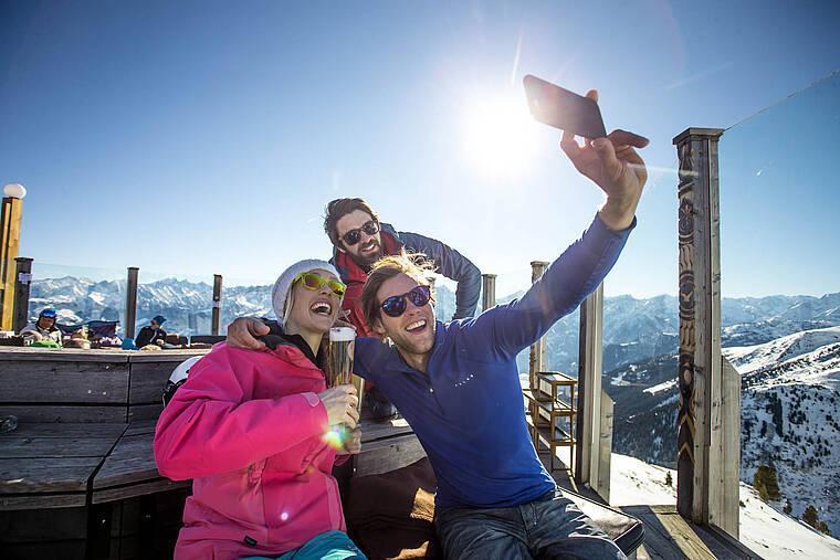 Auf einer Alm im Skigebiet Zillertal sitzen drei Freunde zusammen und machen ein Selfie vor traumhafter Alpenkulisse