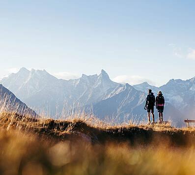 Ein Paar steht bei einem Wanderausflug im Sommerurlaub in Tirol auf einer Almwiese im Sonnenuntergang und genießt das atemberaubende Alpenpanorama