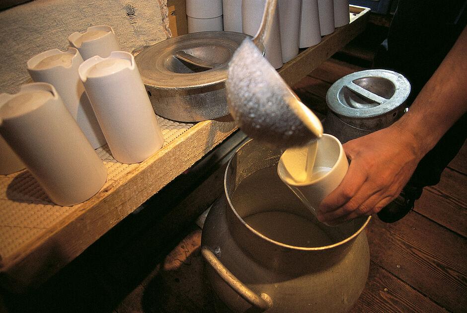 Mit einer Kelle wird aus einer eisernen Milchkanne Milch in kleinere Gefäße abgefüllt