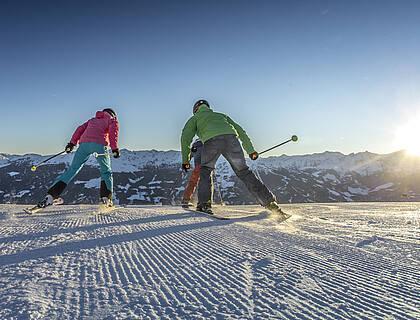 Drei Gäste vom Hotel Kaltenbach nehmen beim Skifahren Schwung, während sie auf eine winterliche Bergkette zusteuern