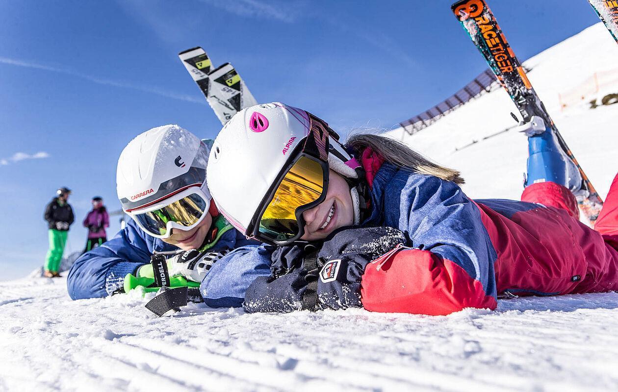 Zwei Kinder in Skiausrüstung liegen im Winterurlaub in Tirol auf dem Bauch im Schnee und lächeln in die Kamera