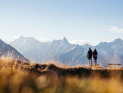 Ein Paar steht bei einem Wanderausflug im Sommerurlaub in Tirol auf einer Almwiese im Sonnenuntergang und genießt das atemberaubende Alpenpanorama