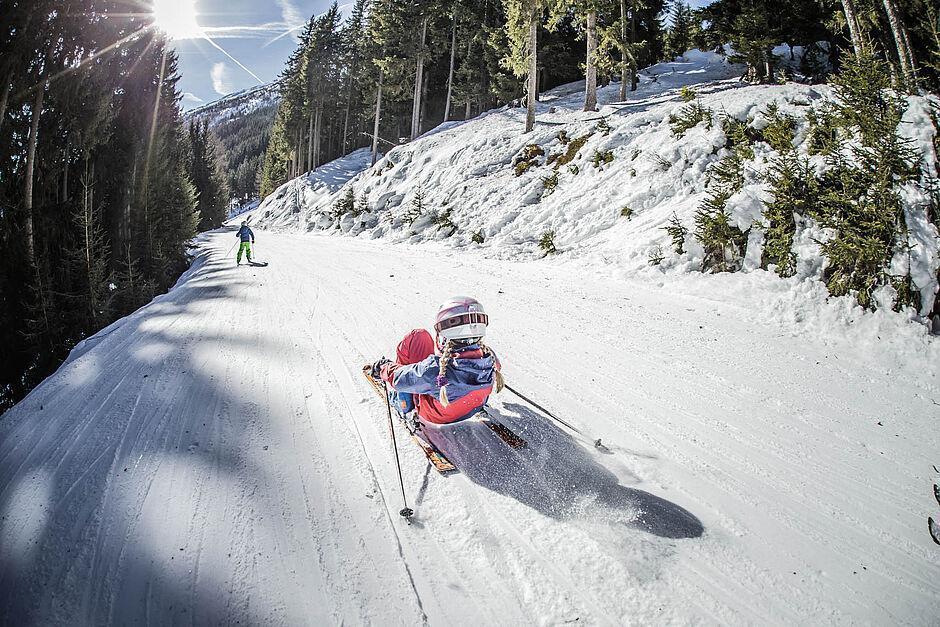 Ein Mädchen fährt wagemutig in der Hocke eine präparierte Piste entlang, die durch den dichten Wald führt