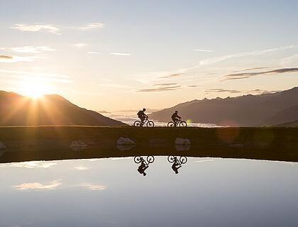 Zwei Fahrradfahrer fahren in der Abendstimmung an einem See in den Bergen