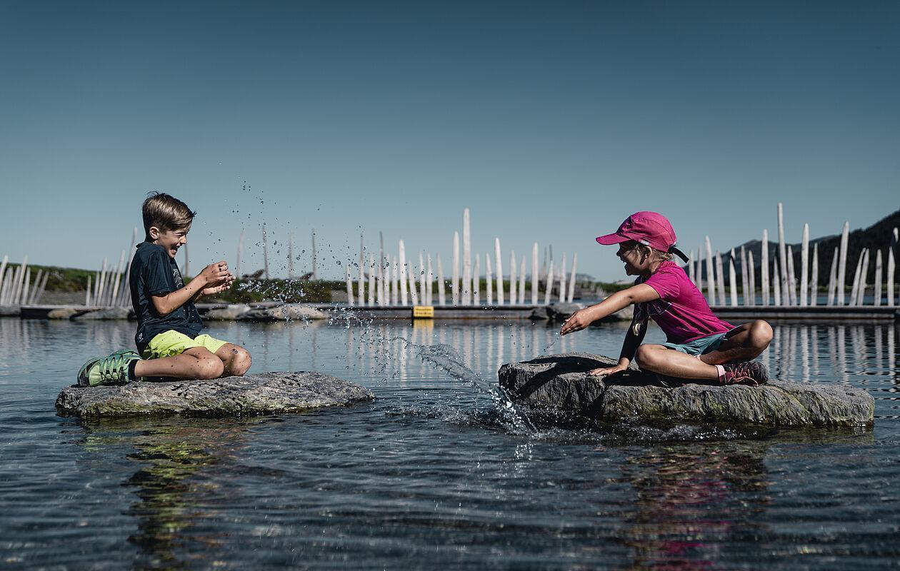 Kinder spielen im Familienurlaub auf zwei großen Steinen im See 